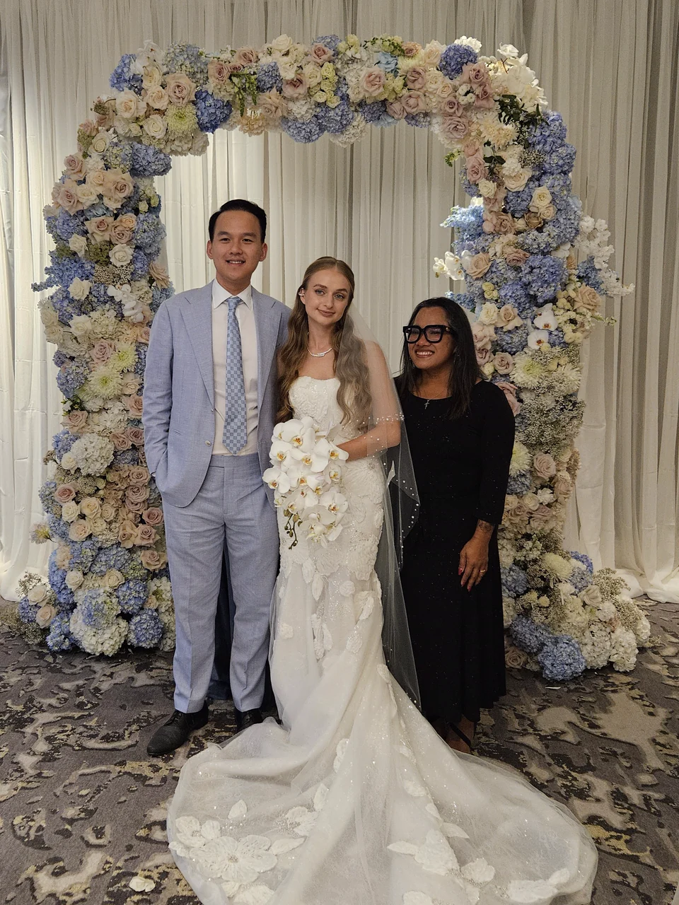 Hong Tam standing with a bride and groom beneath a blue and ivory floral arch after their bilingual wedding ceremony